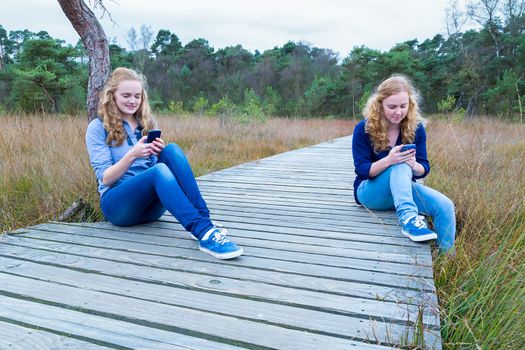 Two caucasian teenage girls operating mobile phones on wooden path in nature