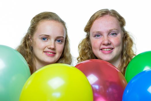 Two smiling caucasian teenage girls behind various colored balloons isolated on white background