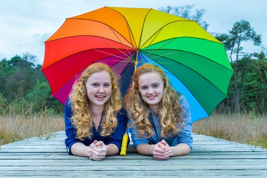 Two smiling caucasian teenage girlfriends lying in nature under colorful umbrella