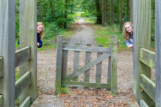 Two caucasian teenage girls hiding at wooden entry of hiking trail in nature