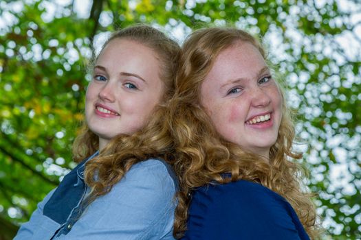 Two caucasian teenage sisters heads together laughing under green tree