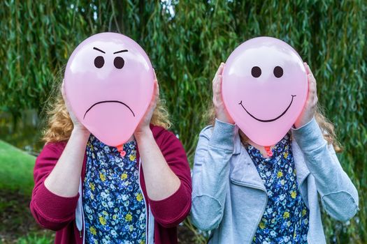 Two teenage girls holding balloons with smiling and angry facial expressions outdoors