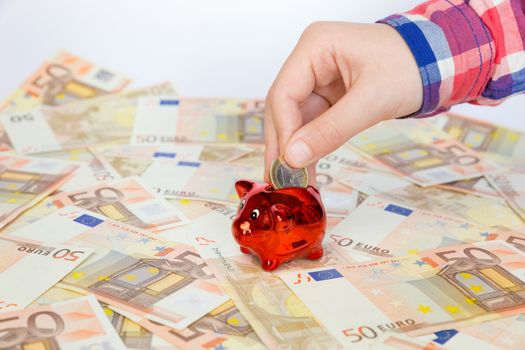 Hand of child putting euro coin in red piggy bank on many spread euro notes
