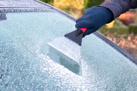 Hand wearing glove scratching ice from car window with frost in winter