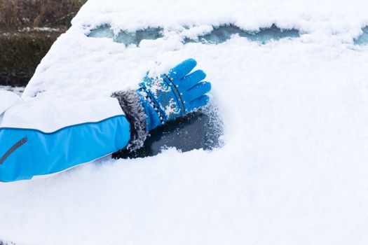 Arm with blue glove removing snow from car windowin winter season
