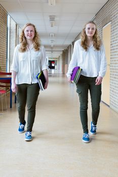 Two caucasian teenage girls walking in long school corridor carrying text books