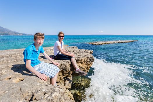 Mother and son as tourists sitting on rock at blue sea enjoying vacation