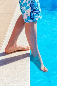 Legs of teenage boy with foot feeling water temparature in swimming pool on sunny day in summer