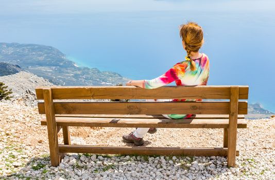 Middle aged woman sitting on brown wooden bench on top of mountain enjoying vacation in Greece