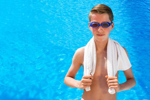 European teenage boy wearing swimming goggles and white towel at blue swimming pool