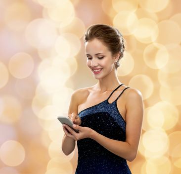 technology, communication and people concept - smiling woman in evening dress holding smartphone over holidays lights background