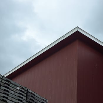 Side of a red building with contrasting roof edge against an overcast sky near a stack of empty skids.