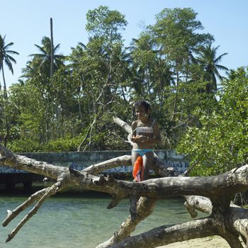A yound girl from the tropics sits on a tree over water in a lush location 