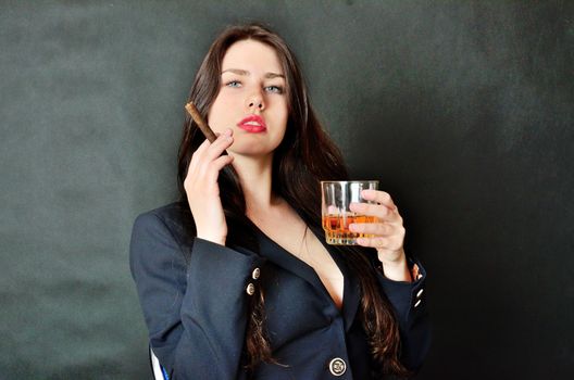 Pretty brunette model holding glass of whisky and cigar. Girl with confident face expression, wearing dark tail-coat.