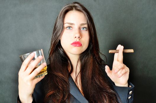Pretty brunette model holding glass of whisky and cigar. Girl with confident face expression, wearing dark tail-coat.