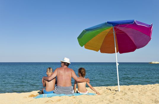 Grandfather hugging his grandson and granddaughter on a beach, looking to the see, back to the camera, colorful umbrella next to them.