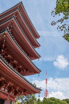 A 5 story Japanese pagoda positioned next to a modern red and white radio tower and a blue sky with clouds in the background.