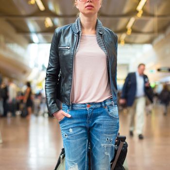 Casually dressed young stylish female traveller walking the airport terminal hall   draging suitcase behind her. Blured background. Can also be used as railway, metro, bus station.