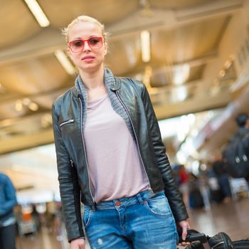 Casually dressed young stylish female traveller walking the airport terminal hall   draging suitcase and a handbag behind her. Blured background. Can also be used as railway, metro, bus station.