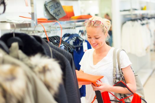 Woman shopping clothes. Shopper looking at clothing indoors in store. Beautiful blonde caucasian female model wearing casual clothes and fashionable sunglasses. Focus on model, shallow depth of field.