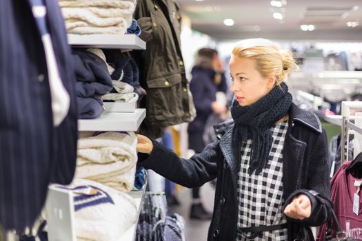 Woman shopping clothes. Shopper looking at clothing indoors in store. Beautiful blonde caucasian female model wearing casual winter clothing and black scarf.