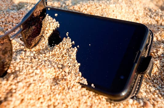 Smart phone and sunglasses buried in the sand. Concept for summer vacation on the beach.