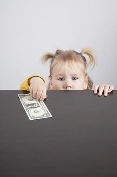 portrait of blonde caucasian baby nineteen month age with pigtails chubby face yellow shirt looking at camera and taking a dollar banknote on brown table