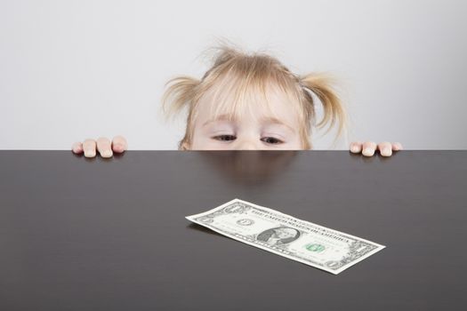 portrait of blonde caucasian baby nineteen month age with pigtails chubby face yellow shirt looking at dollar banknote on brown table horizontal