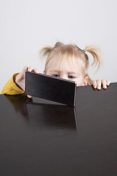 portrait of blonde caucasian baby nineteen month age chubby face yellow shirt watching mobile phone smartphone with her hand on brown table