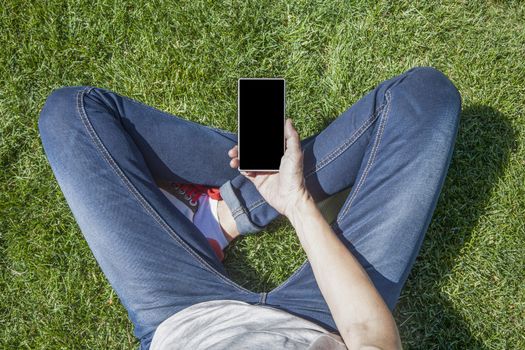 hand of woman blue jeans trousers legged with mobile phone smartphone blank screen sitting on green grass lawn in park