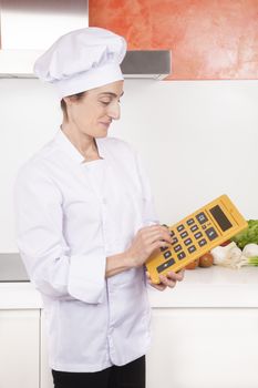 portrait of brunette happy chef woman with professional jacket and hat in white and orange kitchen typing screen big yellow calculator with large numbers