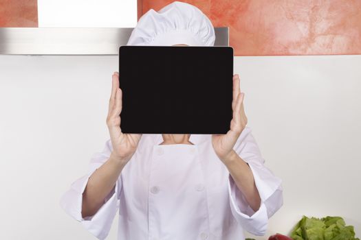 portrait of brunette happy chef woman with professional jacket and hat in white and orange kitchen showing blank screen tablet horizontal