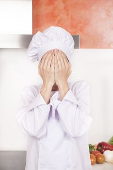 portrait of brunette chef woman with professional jacket and hat in white and orange kitchen hidden face with both hands