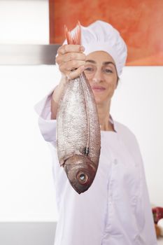 portrait of brunette happy chef woman with professional jacket and hat  in white and orange kitchen with raw fresh fish sea bream in her hand