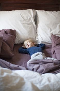portrait of happy blonde caucasian baby nineteen month age with blue shirt sleeping in brown sheets bed between cushions