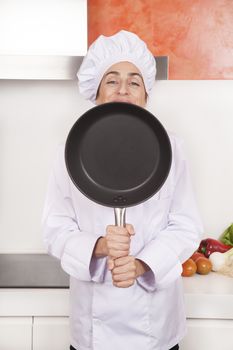 portrait of happy brunette chef woman with professional jacket and hat in white and orange kitchen hidden face looking over steel blank pan