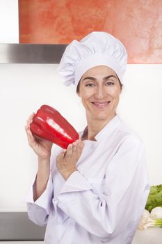 portrait of brunette happy chef woman with professional jacket and hat  in white and orange kitchen with raw fresh big red pepper in her hands