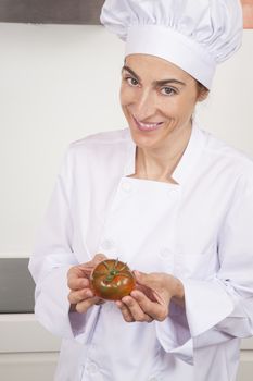 portrait of brunette happy chef woman with professional jacket and hat  in white and orange kitchen with raw fresh red tomato in her hands