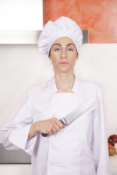 portrait of serious brunette chef woman with professional jacket and hat in white and orange kitchen saluting oath with big metal knife on chest