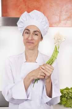 portrait of brunette happy chef woman with professional jacket and hat  in white and orange kitchen with bunch of raw natural garlic shoots in her hands
