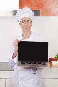 portrait of brunette happy chef woman with professional jacket and hat in white and orange kitchen showing blank screen laptop computer pc