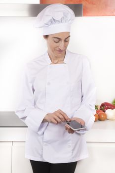 portrait of brunette happy chef woman with professional jacket and hat in white and orange kitchen using blank screen mobile phone