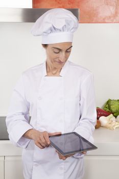 portrait of brunette happy chef woman with professional jacket and hat in white and orange kitchen using blank screen tablet