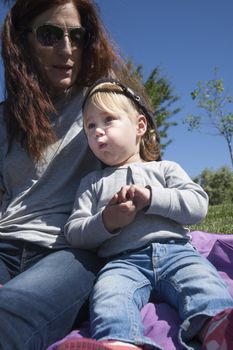 portrait of two years age blonde baby with black cap eating banana next to woman mother in park