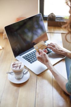 woman with green shirt typing on keyboard pc laptop with made up credit card in hand next to cappuccino coffee cup on light brown wooden table