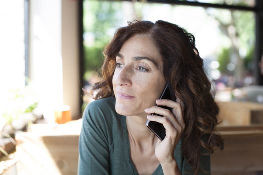 portrait of woman with green shirt curly brunette hair with listen to mobile phone in ear sitting inside light brown wooden cafe
