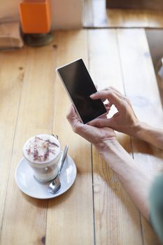 woman hands touching mobile phone with cappuccino coffee cup on light brown wooden table cafe