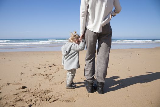 two years age blonde baby with grey coat and brunette mother woman holding hands looking at ocean in sand of beach in front of water sea or ocean