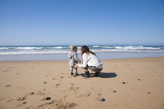 two years age blonde baby with grey coat and brunette mother woman squatting in sand of beach in front of water sea or ocean