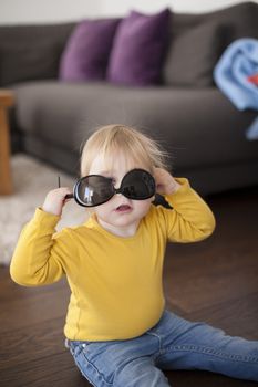 two years aged blonde happy baby yellow shirt and blue jeans playing with black sunglasses of woman mother sitting on wooden indoor floor and looking at camera